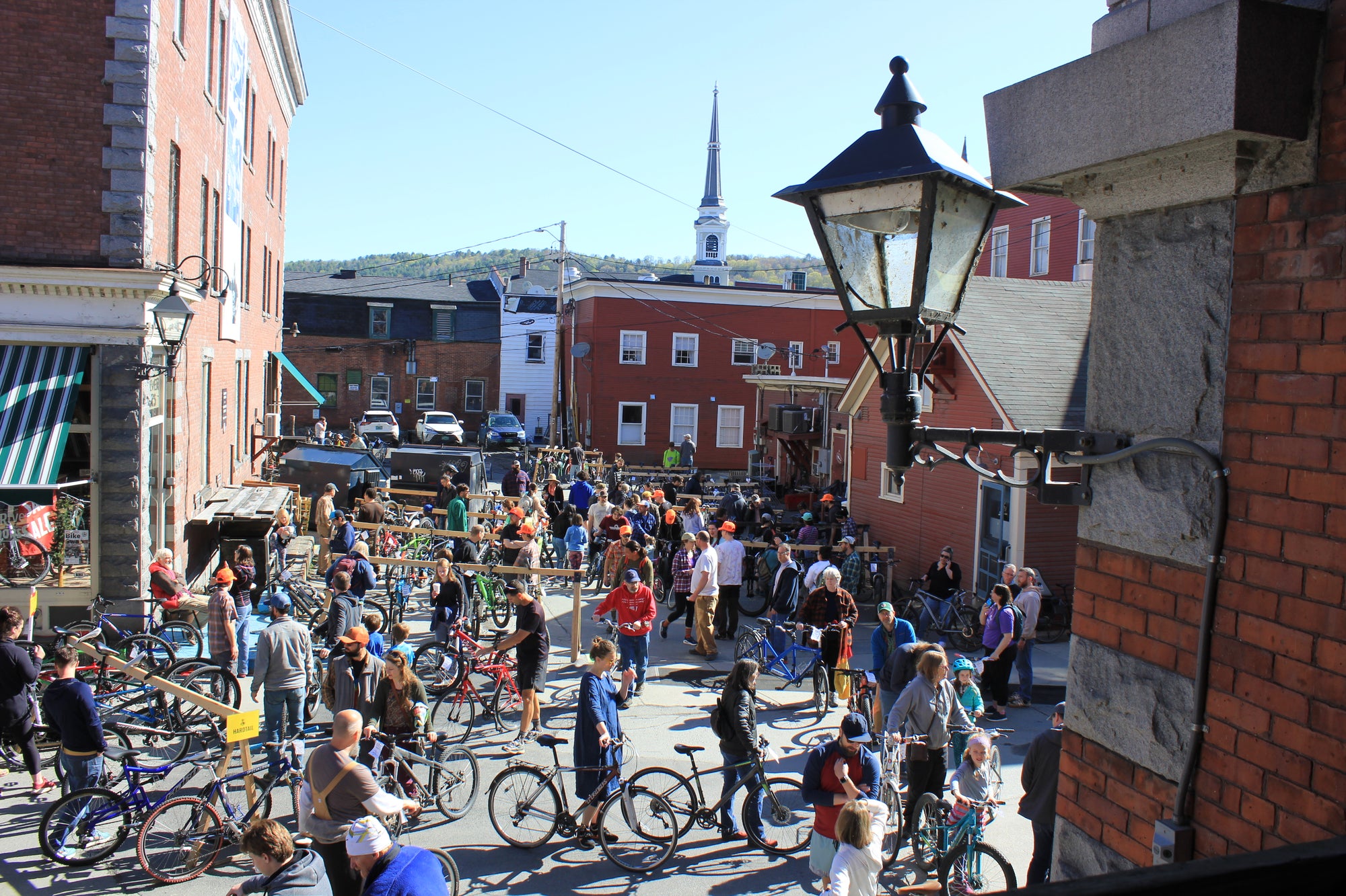 People walking around looking at used bikes resting on wooden racks in between brick buildings 