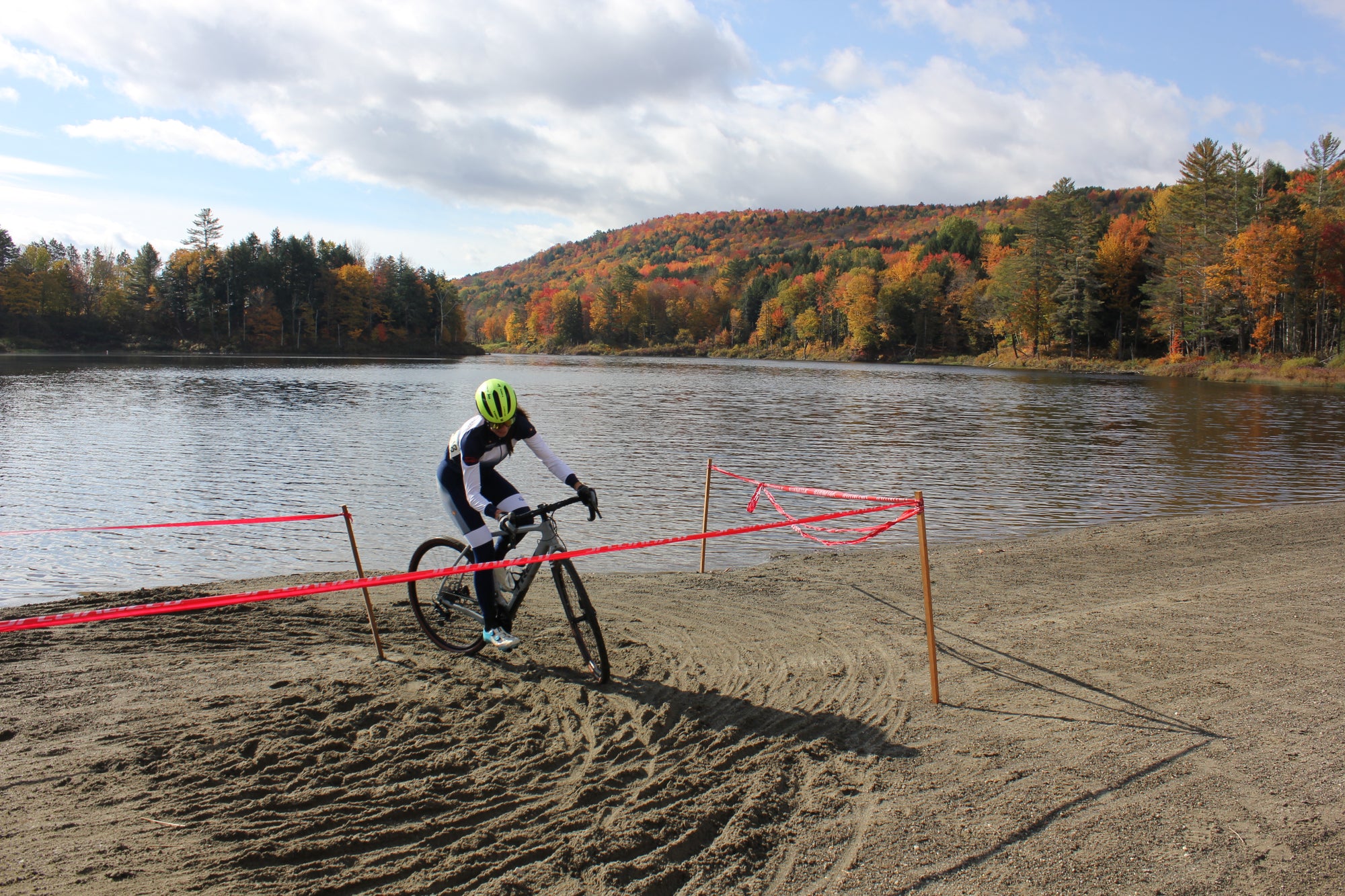 A bike rider racing cyclocross on a beach with a lake behind them