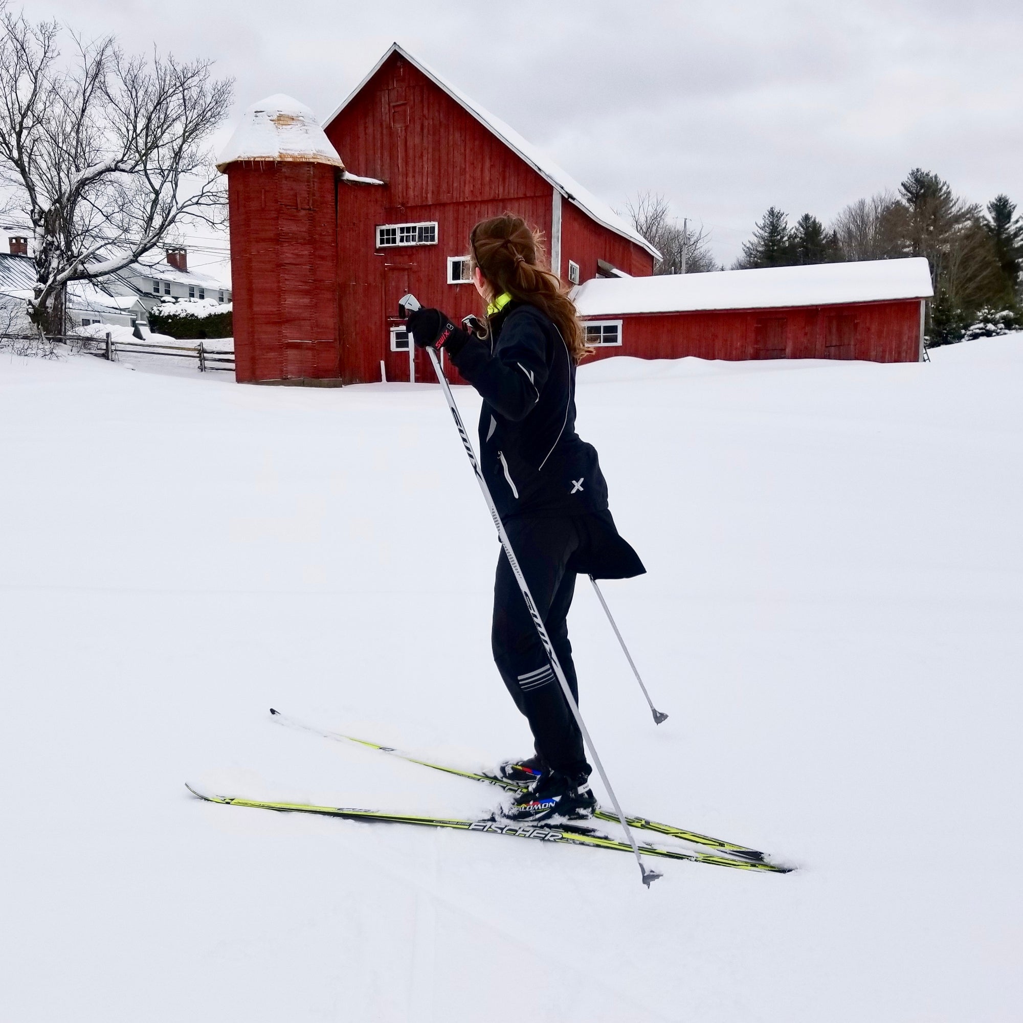A cross country skier dressed in black with yellow skis looking at a red barn