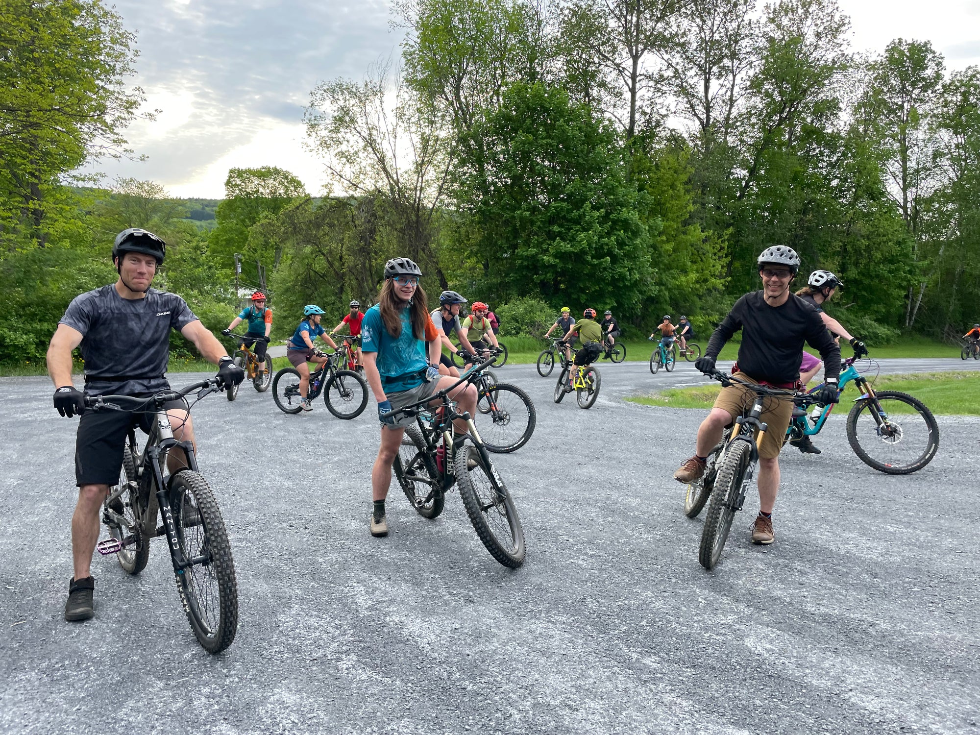 three smiling mountain bikers with their bicycles and a group of riders milling around behind them