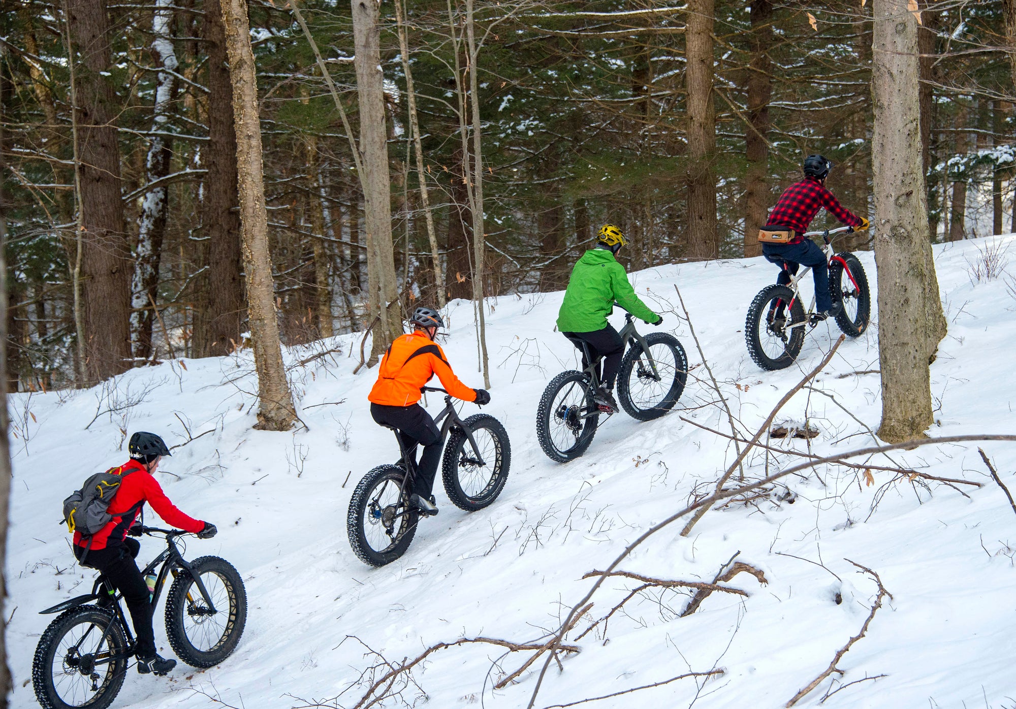 Four fat bikers in red, orange, green and plaid jackets riding on a snowy trail
