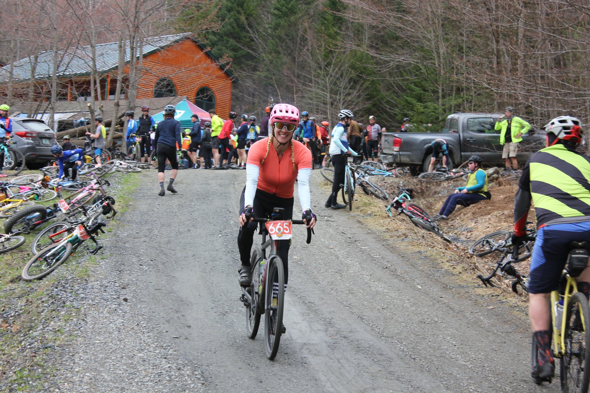 A cyclist in an orange jersey riding down a dirt driveway away from a group of people