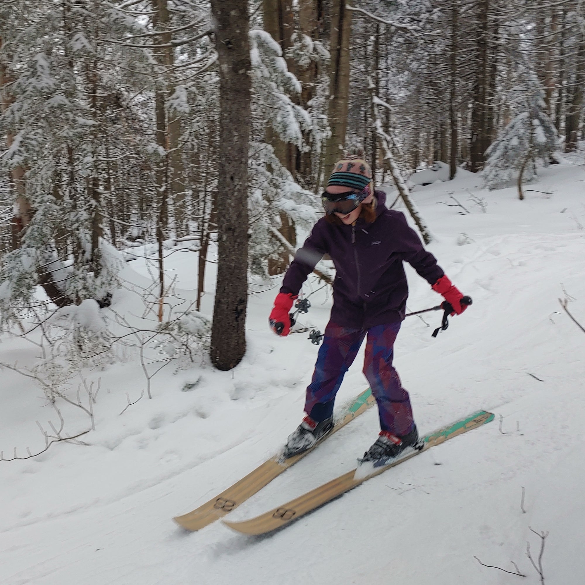 A skier in a purple jacket and pink mittens skiing downhill through the woods on altai skis