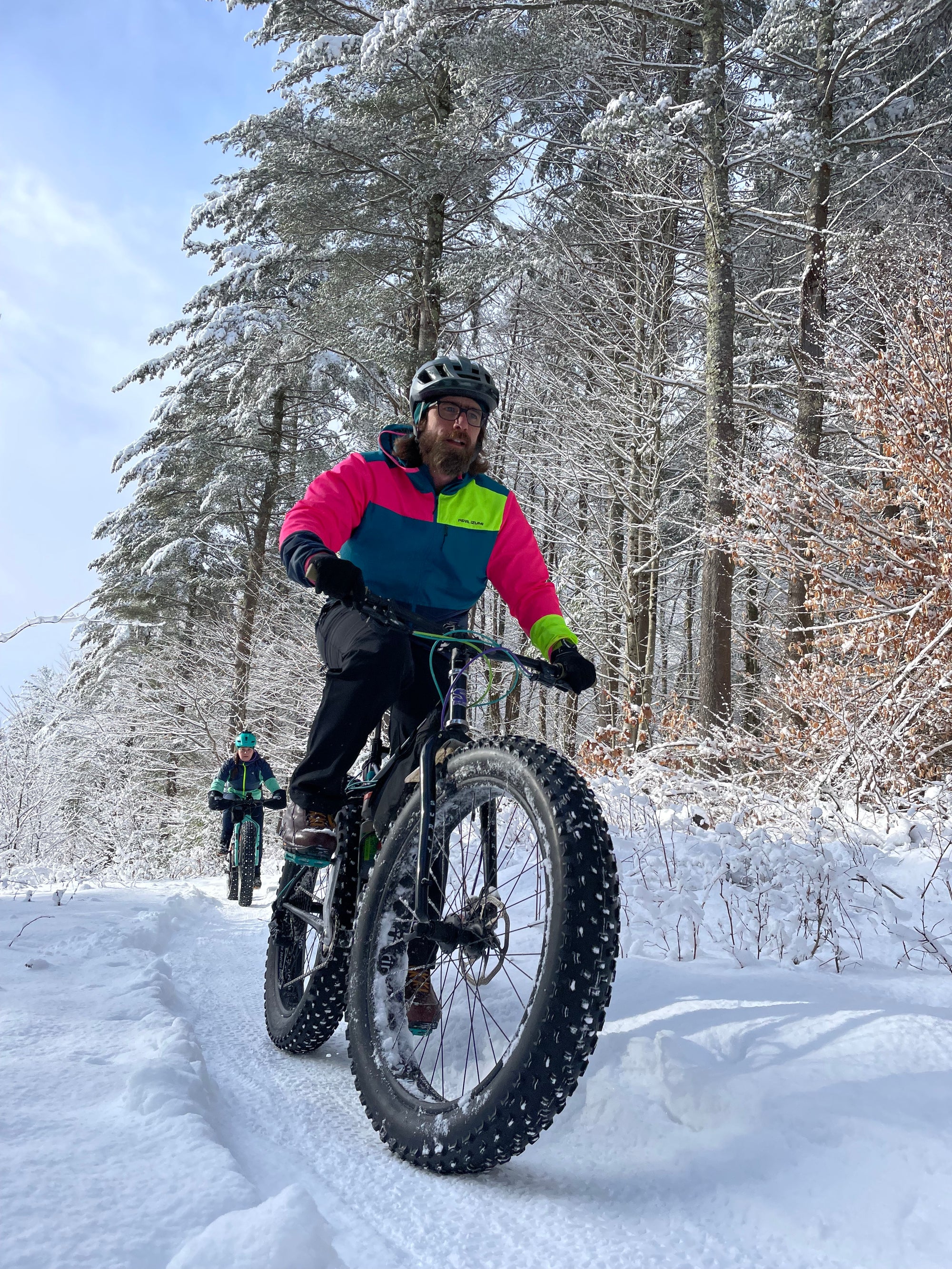 A fat biker in a bright pink, yellow and teal jacket riding on a packed snowy trail