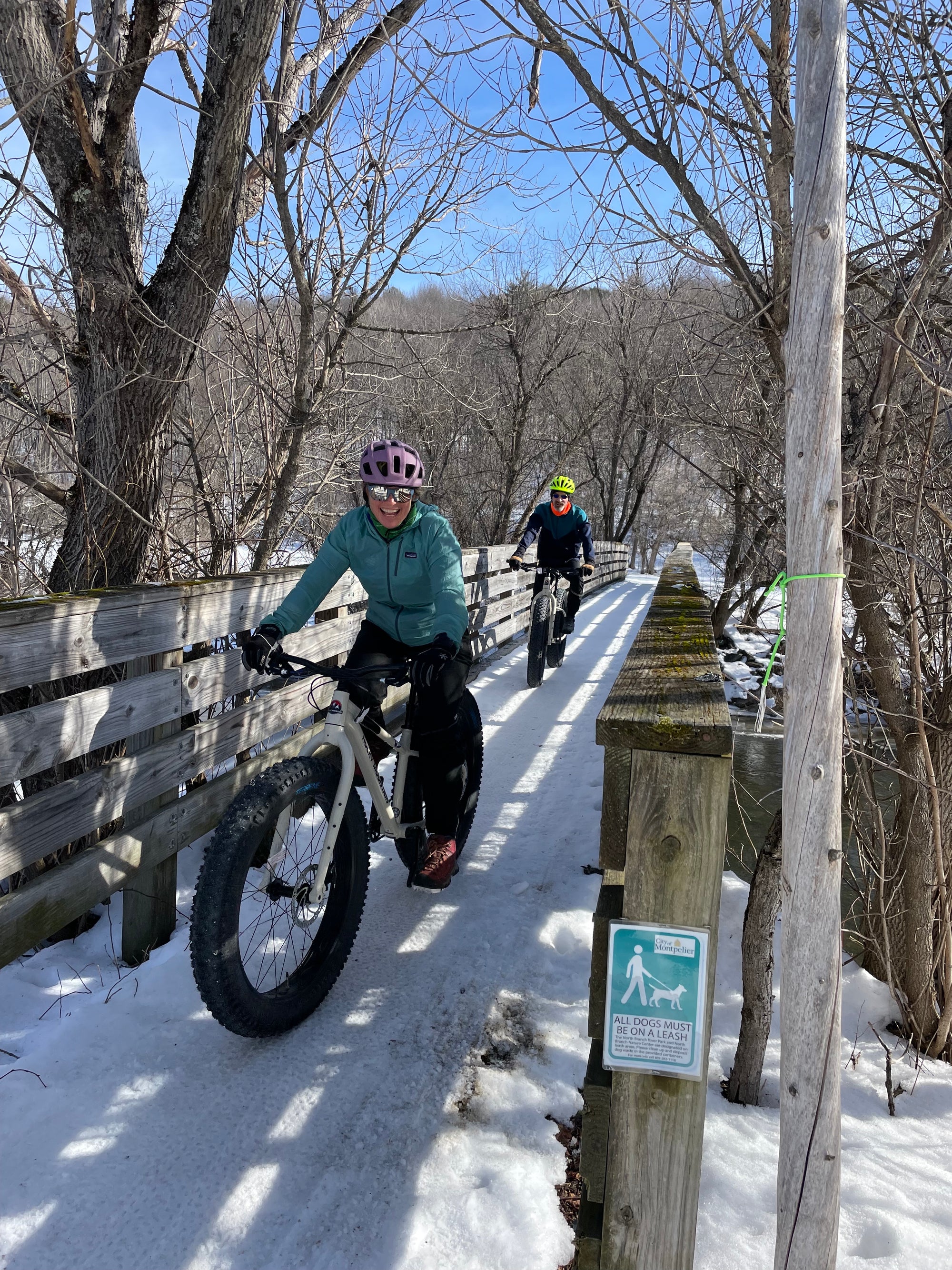 Two fat bikers riding single file on a wooden bridge in late winter